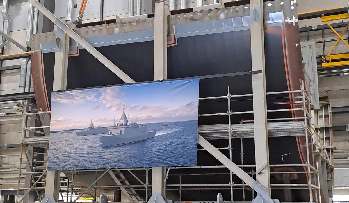 The image shows the steel keel section of the multi-role corvette, with a large illustrated rendering of the completed vessel displayed in front of it.