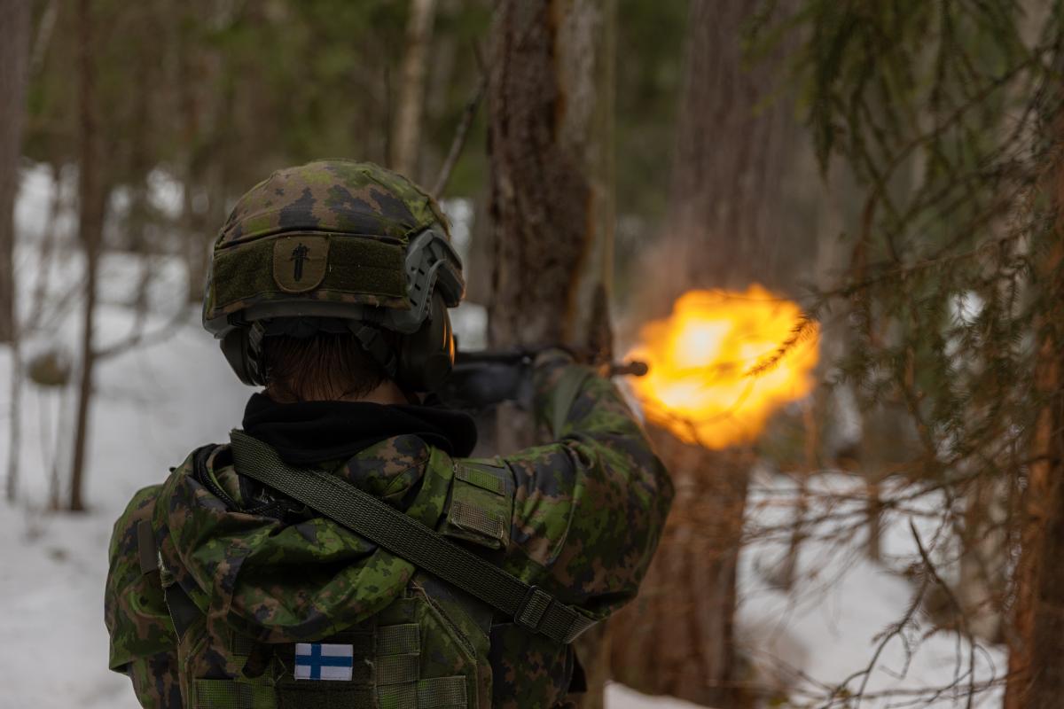 Sotilaspoliisi ampuu rynnäkkökiväärillä lumisessa metsässä. Rynnäkökivääristä tulee suuliekki. Sotilas on selin kameraan päin.