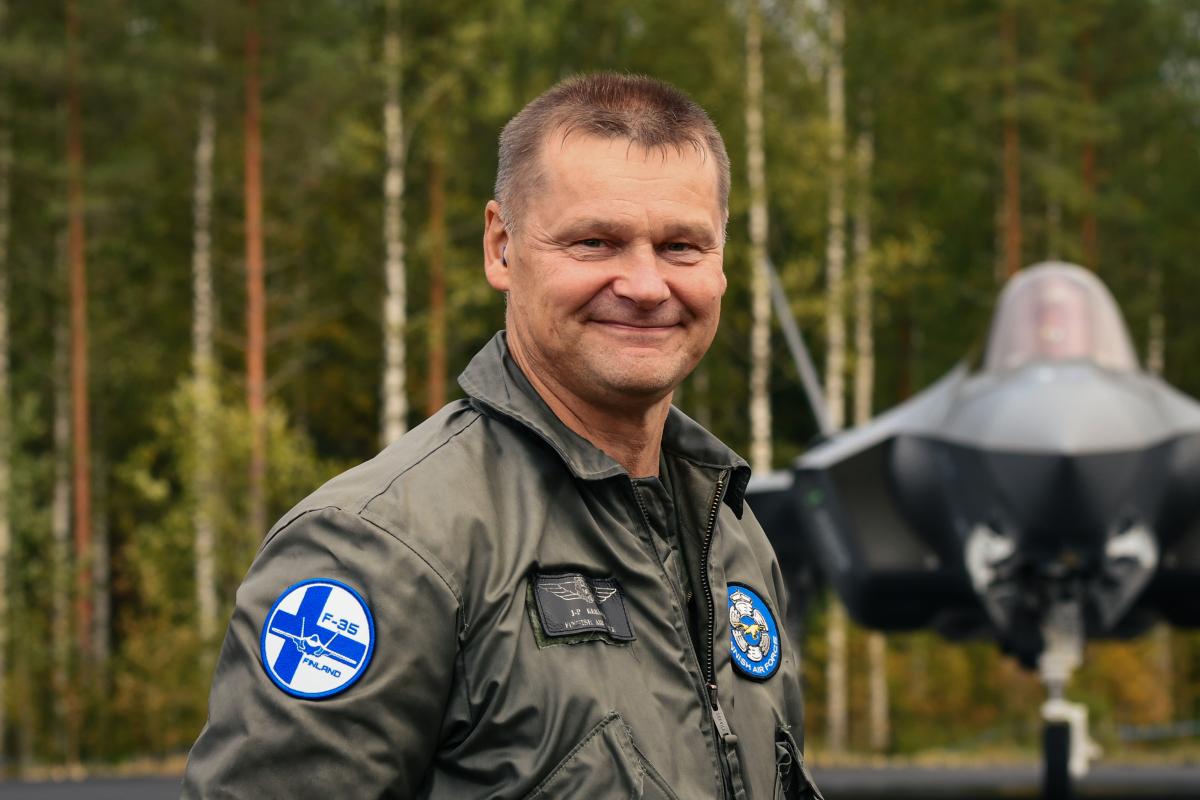 Kenraalimajuri Keränen katsoo kameraan ja hymyilee. Hän on pukeutunut lentohaalariin, jonka hihassa on F-35-hankkeen hihamerkki. Keräsen taustalla näkyy norjalainen F-35-hävittäjä pysähtyneenä asfaltilla. Taaimmaisena on syksyistä suomalaista sekametsää.