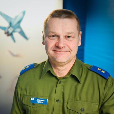 Lähikuvassa vihreään kauluspaitaan pukeutunut kenraalimajuri Keränen katsoo kameraan ja hymyilee. Taustalla näkyy taulu Hornet-hävittäjästä.