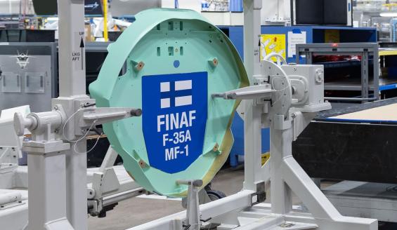 Tehtaalla on harmaassa eräänlaisessa telineessä kiinni turkoosin sininen, hieman lautasen mallinen esine. Esine on osa Suomen ensimmäisen F-35-hävittäjän eturunkoa. Osaan on kiinnitetty laatta, jossa on Suomen lippu ja lukee FINAF ja F-35-A, eli Suomen ilmavoimien F-35A.