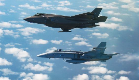 Alankomaalainen F-35 ja suomalainen Hornet lentävät vierekkäin, kuvakulman vuoksi lähes päällekkäin, sinisellä, rakoilevan pilvisellä taivaalla.